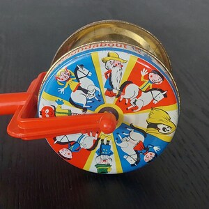 May include: Vintage toy with a red handle and a colorful, rotating drum. The drum features illustrations of children, horses, and a bearded man. The words "Roundabout" are printed around the drum's edge. The toy is made of metal and plastic.