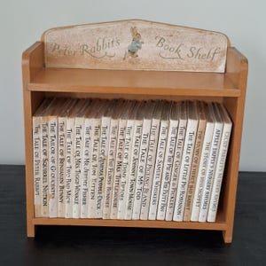 May include: A light brown wooden bookshelf with a decorative top panel featuring an illustration of Peter Rabbit and the text Peter Rabbit's Book Shelf. The shelf holds a collection of twenty-three small books with white spines and black text.