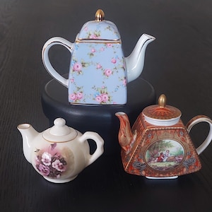 Peut inclure: Trois théières décoratives. La théière supérieure est bleu clair avec un motif floral et des accents dorés. La théière du milieu est blanche avec un motif floral. La théière du bas est orange avec une scène de paysage.