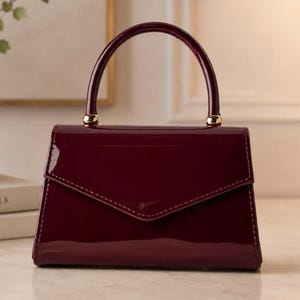 May include: A structured handbag in a deep burgundy patent leather finish with a glossy sheen. The bag features a triangular envelope-style flap closure, a single rounded top handle, and polished gold-tone hardware accents at the handle base.
