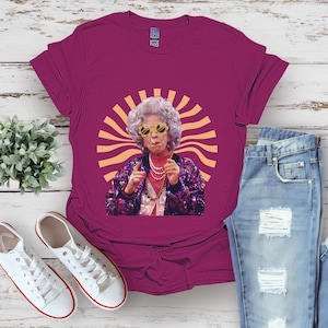 May include: A magenta t-shirt with a graphic of a woman wearing sunglasses and a sparkly outfit. The woman is standing in front of a yellow sunburst.
