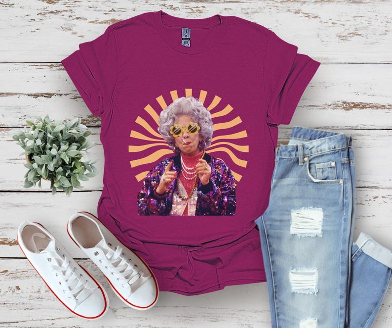 May include: A magenta t-shirt with a graphic of a woman wearing sunglasses and a sparkly outfit. The woman is standing in front of a yellow sunburst.