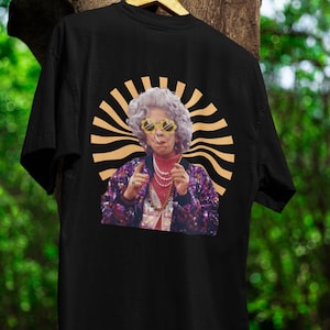 May include: Black t-shirt with a graphic print of a woman wearing sunglasses and a sequined jacket. The graphic is surrounded by a gold sunburst design.