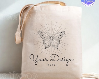 Op de afbeelding: Beige canvas draagtas met een zwart-wit vlinderontwerp en de tekst "Your Design Here". De vlinder is omgeven door stralende lijnen. De tas heeft lange handvatten.