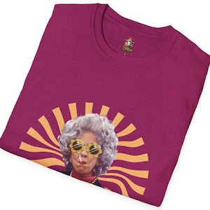 May include: A magenta t-shirt with a graphic of a woman wearing yellow sunglasses and a cigarette. The woman has gray hair and is surrounded by a yellow sunburst design.