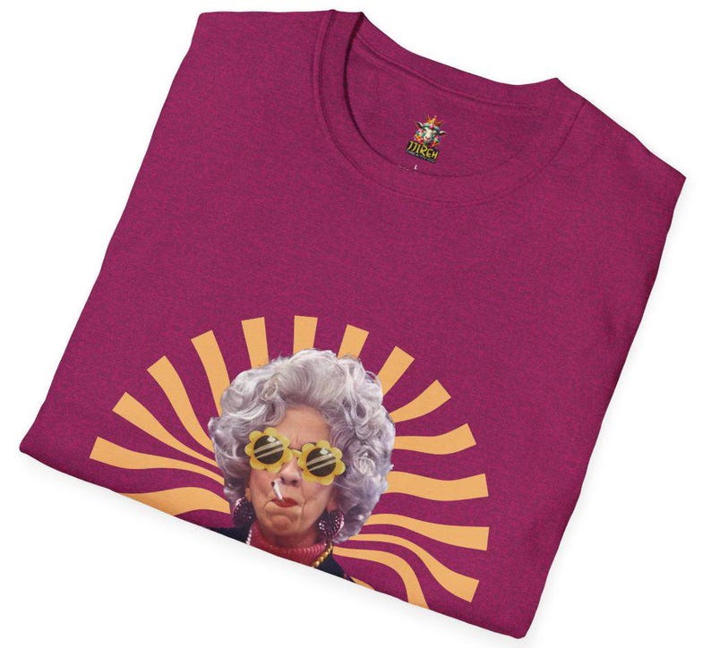 May include: A magenta t-shirt with a graphic of a woman wearing yellow sunglasses and a cigarette. The woman has gray hair and is surrounded by a yellow sunburst design.