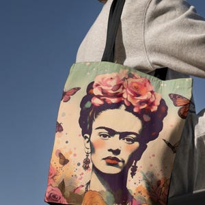 Könnte beinhalten: Eine Tragetasche mit einem Porträt von Frida Kahlo mit floralem Kopfschmuck und Schmetterlingsakzenten. Die Tasche hat einen hellgrünen Hintergrund mit rosa Rosen und Schmetterlingen. Die Tragetasche wird vor blauem Himmel gehalten.