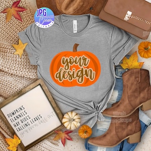 May include: A grey t-shirt featuring an orange pumpkin graphic with the text "your design." The image includes autumn-themed accessories such as a brown handbag, boots, and fallen leaves. A framed sign lists autumn-related words.