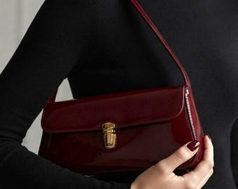 May include: A rectangular shoulder bag in a deep burgundy patent leather finish. The bag features a smooth, glossy surface and a gold-toned metal push-lock closure centered on the front flap. It includes a thin, matching burgundy shoulder strap.
