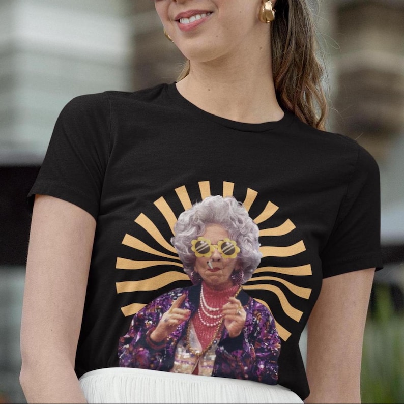 May include: Black t-shirt with a graphic of a woman wearing sunglasses and a sequined jacket. The graphic is surrounded by a yellow and orange sunburst design.