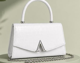 May include: A white handbag with a crocodile-embossed texture, featuring a single top handle and a silver-toned V-shaped metal emblem on the front flap. The bag includes a detachable silver-toned chain shoulder strap and a structured rectangular shape.