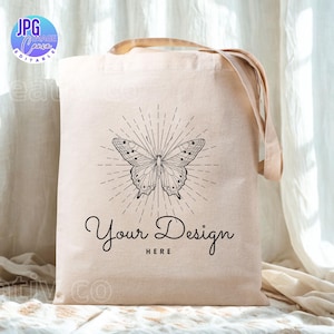 May include: A beige canvas tote bag featuring a black and white butterfly design with radiating lines. The text "Your Design Here" is below the butterfly. The bag has beige handles and is set against a neutral backdrop.