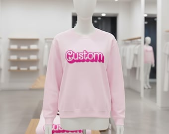 May include: A light pink sweatshirt with the word "Custom" in a magenta, bubble-style font. The sweatshirt is displayed on a mannequin in a retail setting, with folded sweatshirts visible below.