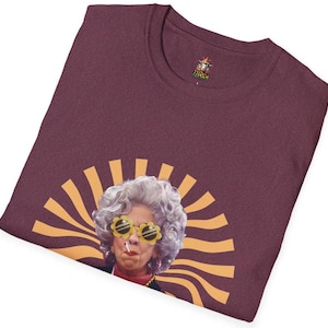 May include: A maroon t-shirt with a graphic of a woman with gray hair wearing yellow sunglasses and a cigarette. The woman is in front of a yellow and orange sunburst design.