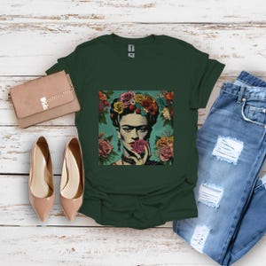 May include: A dark green t-shirt with a colorful graphic print of Frida Kahlo wearing a flower crown and holding a flower. The graphic is on a light blue background.
