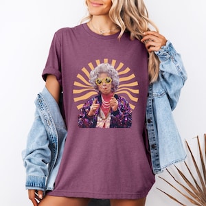May include: A purple t-shirt with a retro-style graphic of a woman wearing sunglasses and a sequined jacket. The graphic is surrounded by a yellow sunburst design.