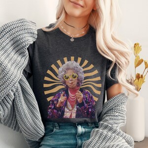 May include: A dark gray t-shirt with a graphic of a woman wearing sunglasses and a sequined jacket. The graphic is set against a yellow and brown sunburst background.