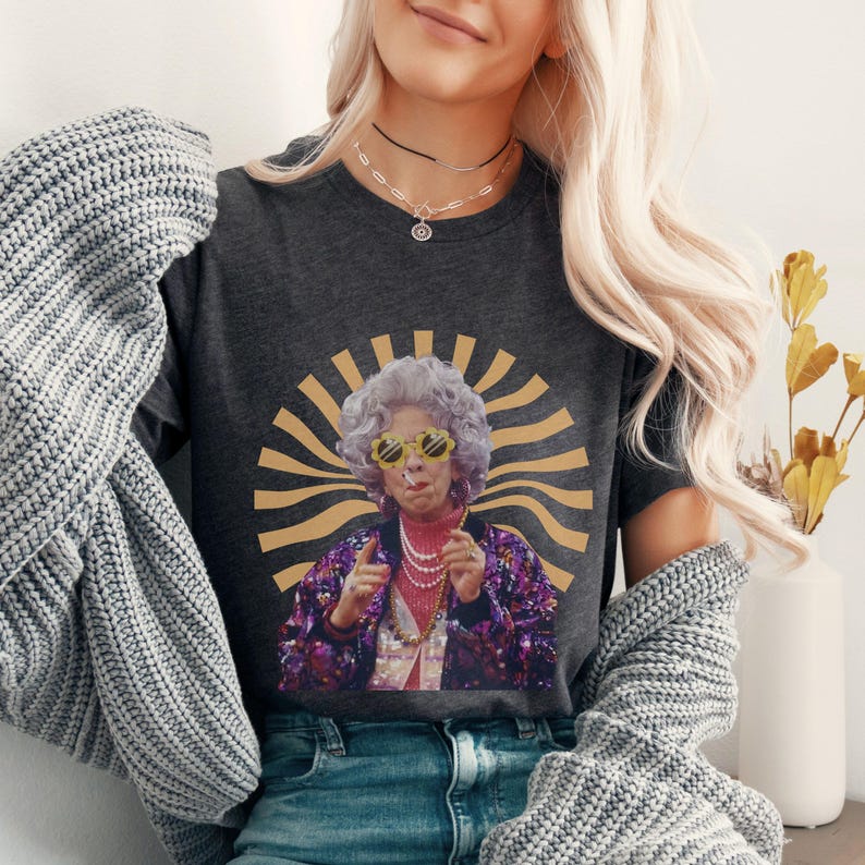May include: A dark gray t-shirt with a graphic of a woman wearing sunglasses and a sequined jacket. The graphic is set against a yellow and brown sunburst background.