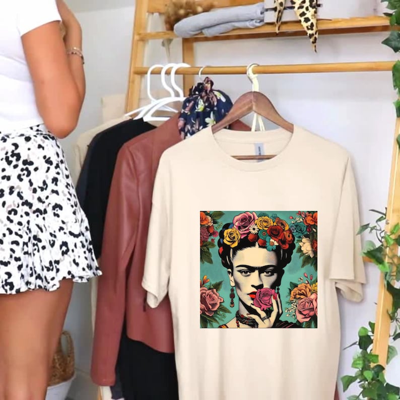 May include: A cream colored t-shirt with a colorful graphic of Frida Kahlo wearing a flower crown and holding a pink flower to her face.