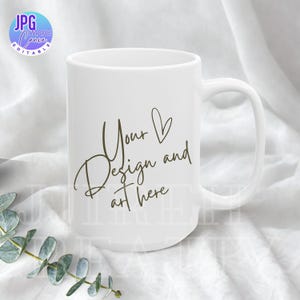 Puede incluir: Una taza de cerámica blanca con un asa curvada. La taza presenta el texto "Your Design and art here" en un estilo manuscrito. La taza está colocada sobre un fondo de tela blanca con una ramita de hojas verdes.