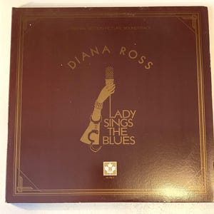 May include: A burgundy vinyl record album cover with gold lettering. The album cover features a woman's hand holding a microphone and a gold handcuff. The album title is "Diana Ross Lady Sings The Blues".
