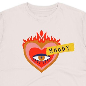 Può includere: Una maglietta bianca con un cuore rosso e rosa con un occhio e fiamme in cima. Il cuore è parzialmente coperto da un nastro giallo con la parola "MOODY" scritta su di esso.