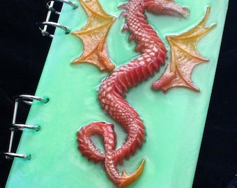 May include: A green spiral bound notebook with a red dragon with yellow wings on the front cover. The text "List" is written on the cover.