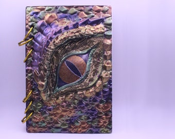 May include: A purple, green, and gold spiral bound journal with a dragon eye embossed on the front cover.