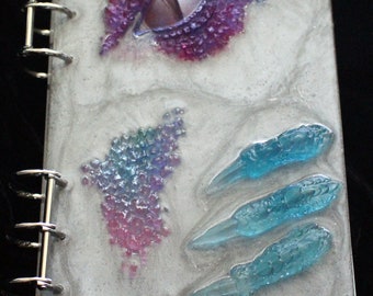 May include: A gray, resin-bound journal with a silver binder. The cover features a purple and blue eye, a cluster of iridescent pink and blue crystals, and three blue claw marks.