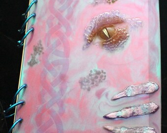 May include: A pink and blue journal with a spiral binding. The cover features a Celtic knot design, a dragon eye, and three dragon claws.
