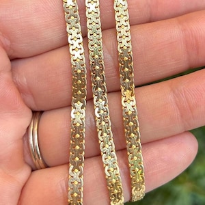 Könnte beinhalten: Nahaufnahme von drei goldfarbenen Halsketten mit einem sich wiederholenden Muster aus ineinandergreifenden Formen. Die Halsketten sind dünn und zart, mit einem Verschluss unten sichtbar. Der Schmuck wird vor einem verschwommenen Hintergrund präsentiert.