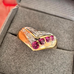 May include: A gold ring with a V-shaped design. The ring features a row of four pink gemstones and is accented with small white gemstones. The ring is set in a gold band and is displayed in a gray jewelry box.