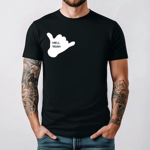 May include: Black t-shirt featuring a white hand gesture graphic with the words "HELL YEAH" inside. The shirt is a classic crew neck style. The person wearing the shirt has tattoos on both arms.