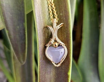 Könnte beinhalten: Ein goldfarbener, drahtgewickelter Anhänger mit einem hellgrauen, herzförmigen Edelstein. Der Anhänger hängt an einer goldenen Kette.