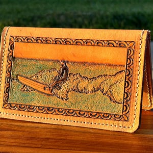 May include: A tan leather wallet featuring a hand-tooled design of a person surfing on a wave. The rectangular wallet has a decorative border of repeating semi-circles and is stitched along the edges with light-colored thread.