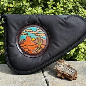 May include: Black canvas pistol case with a leather patch depicting a desert landscape with a cactus and a sunset.