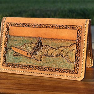 May include: A tan leather wallet with a detailed carving of a surfer on a surfboard. The design features an orange sky and green water, framed by a decorative border. The wallet is open, revealing the interior.