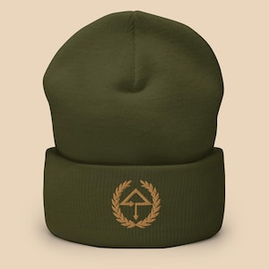 Peut inclure: Un bonnet vert avec un logo brodé marron représentant une flèche pointant vers le haut dans une couronne.