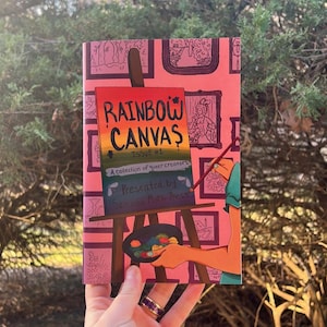 Peut inclure: Une couverture de livre rose avec le titre "RAINBOW CANVAS" aux couleurs de l'arc-en-ciel, avec le texte "ISSUE #1" et "A collection of queer creators". La couverture pr&eacute;sente des illustrations encadr&eacute;es et un artiste peignant sur une toile.