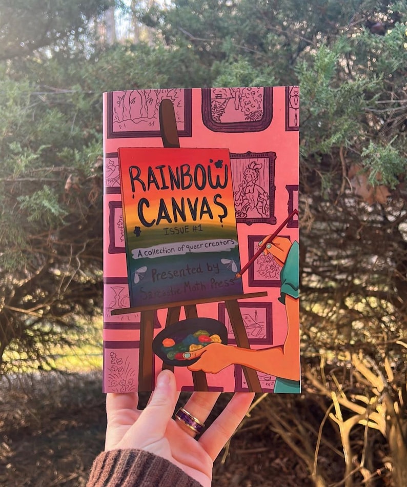 Peut inclure: Une couverture de livre rose avec le titre "RAINBOW CANVAS" aux couleurs de l'arc-en-ciel, avec le texte "ISSUE #1" et "A collection of queer creators". La couverture pr&eacute;sente des illustrations encadr&eacute;es et un artiste peignant sur une toile.
