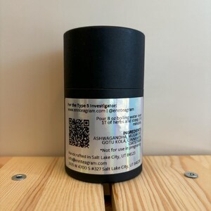 Puede incluir: Un recipiente cil&iacute;ndrico negro con una etiqueta plateada. La etiqueta incluye texto sobre los ingredientes, las instrucciones y el sitio web del producto. El recipiente est&aacute; sobre una superficie de madera.