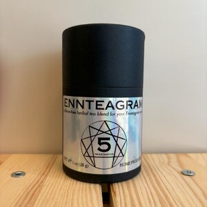 Puede incluir: Un recipiente cil&iacute;ndrico negro de mezcla de t&eacute; de hierbas. La etiqueta dice "ENNTEAGRAM" y "5 INVESTIGATOR". El peso neto es de 28 g. El recipiente est&aacute; sobre una superficie de madera.