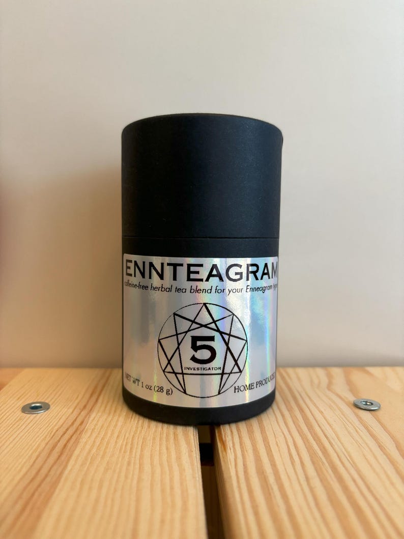 Puede incluir: Un recipiente cil&iacute;ndrico negro de mezcla de t&eacute; de hierbas. La etiqueta dice "ENNTEAGRAM" y "5 INVESTIGATOR". El peso neto es de 28 g. El recipiente est&aacute; sobre una superficie de madera.