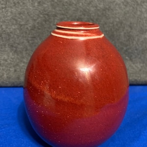 May include: A small, red, egg-shaped ceramic vase with a white rim and two white lines around the top.