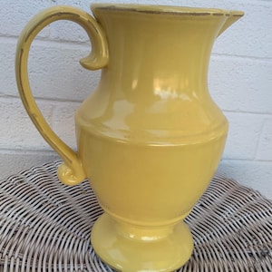 May include: A yellow ceramic pitcher with a wide base and a handle. The pitcher has a smooth, glossy finish.