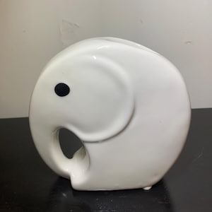 May include: A white ceramic elephant figurine with a black eye. The elephant has a curved trunk and a smooth, glossy finish. The figurine is a decorative item.