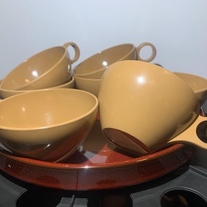 May include: A set of smooth, tan-colored plastic cups with circular handles, arranged on a round, glossy, dark red tray with a speckled finish. The cups feature a wide, open bowl shape and a uniform, solid light brown color throughout the exterior.