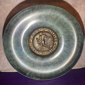 May include: A round, green metal plate with a raised, circular design in the center. The design features a figure of a man in a flowing robe, holding a net, with Hebrew text around the edge.