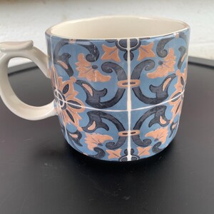 May include: A ceramic mug with a blue, peach, and black floral tile pattern. The mug has a white handle and interior. The design is reminiscent of traditional tile work. The mug is suitable for hot or cold beverages.