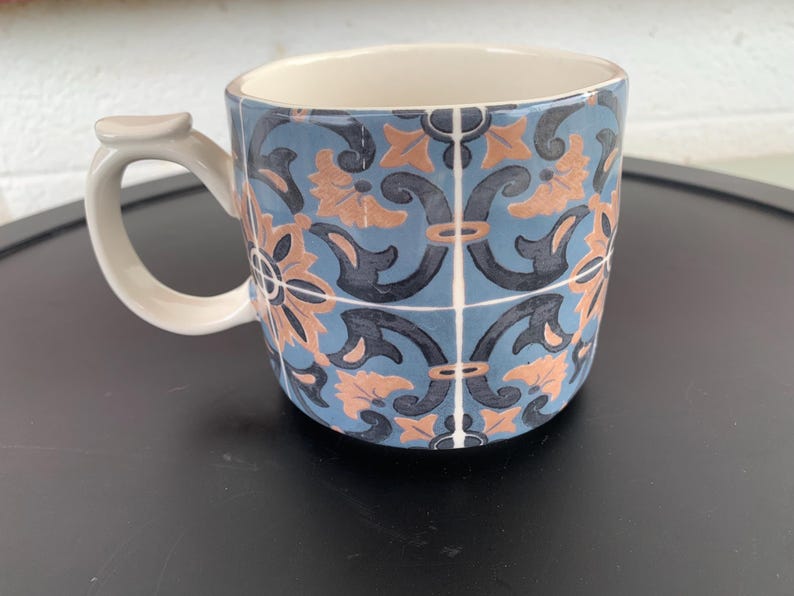 May include: A ceramic mug with a blue, peach, and black floral tile pattern. The mug has a white handle and interior. The design is reminiscent of traditional tile work. The mug is suitable for hot or cold beverages.